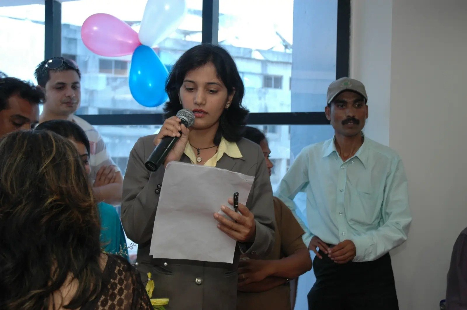 Anjali Wadje speaking at an event, holding a microphone and reading from a paper, with people and balloons in background, candid event photo